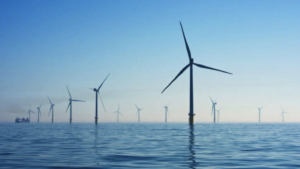A row of wind turbines stands in the ocean, with their blades rotating in the breeze. The scene is set against a clear blue sky, reflecting on the calm water